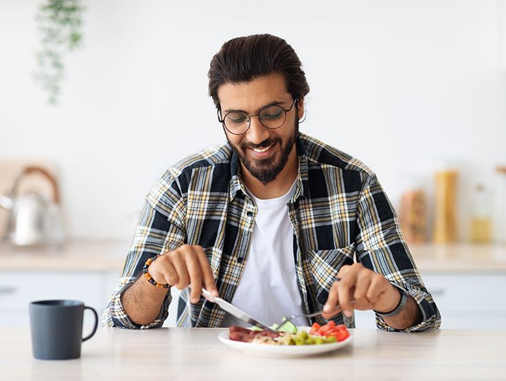 Indian man sitting at a kitchen table eating a healthy meal of vegetables and protein.