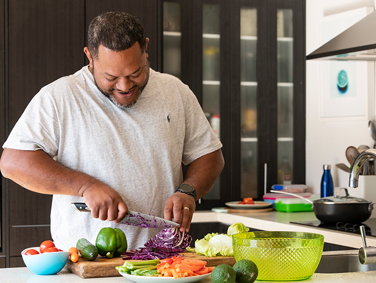 A pasifika man is standing in his kitchen chopping up a wide variety of fresh and colourful vegetables.