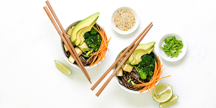 Two Japanese bowls filled with soba noodles, fresh vegetables, avocado and sesame seeds. Also featured are a pair of chop sticks and a fresh lime cut in half.