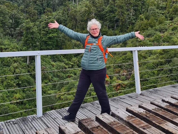 Senior woman wearing hiking clothes standing on a wooden bridge over a river with forest in the background.