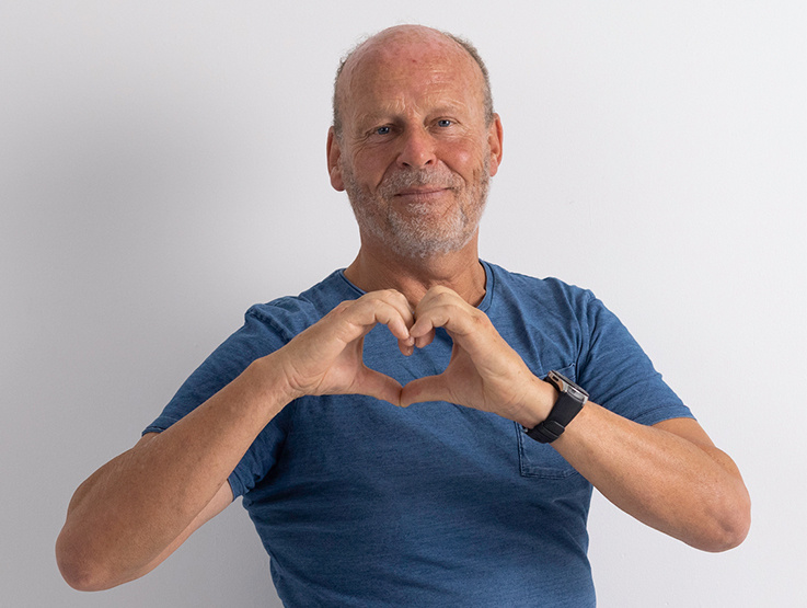 Bruce is an older man in his fifties. He is making the shape of a heart with his hands.