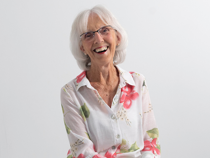 Claire is a senior lady wearing a white and pink floral shirt. She is seated on a stool and looking into the camera smiling.