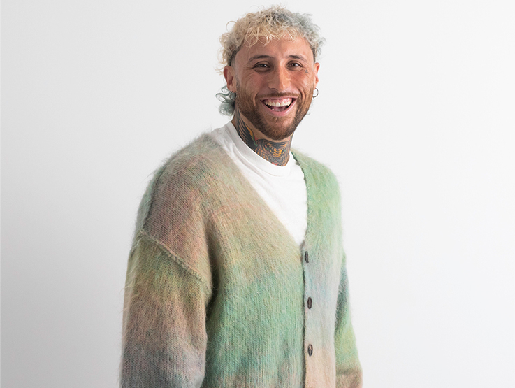 Lance Savali is wearing a pastel fluffy cardigan and is turned towards the camera, laughing.