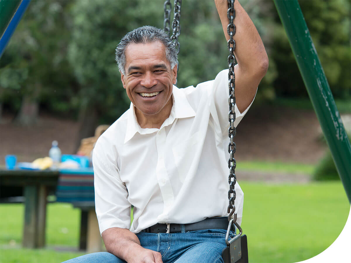 An older Maori man sitting on a swing in a playground with his grandchilden