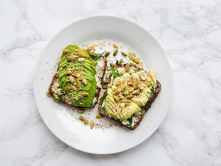 Two pieces of wholegrain toast topped with avocado and nuts and seeds on a white place and a white and gray marble surface.